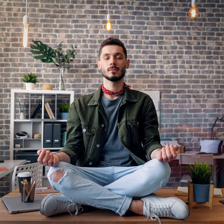 Un jeune homme assis en tailleur sur son bureau en posture de méditation (yoga) pour soulager les maux de dos au travail.